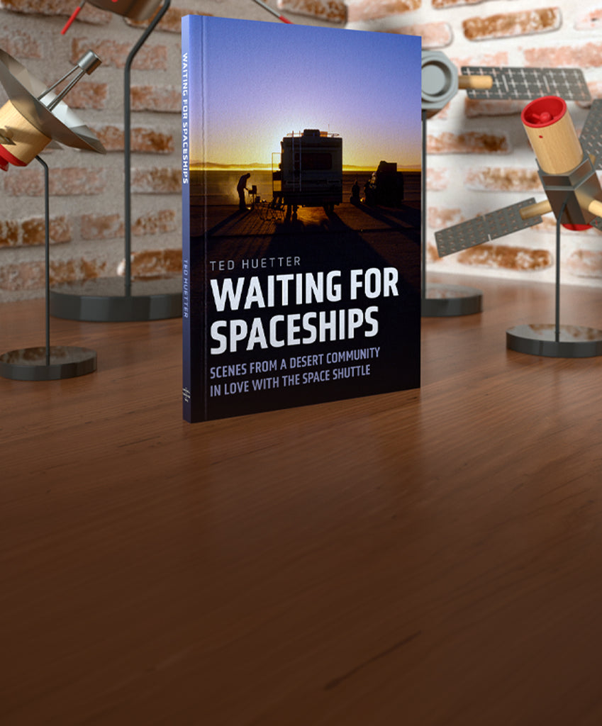 Book titled 'Waiting for Spaceships' by Ted Huettner on a wooden surface with model rocket launchers in the background.