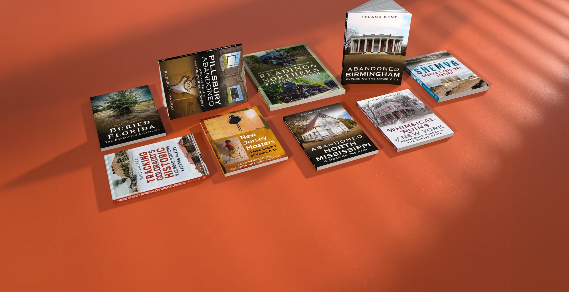A set of nine travel guide books arranged on a warm orange surface with diagonal shadows. The books feature various cover designs with titles such as 'Abandoned Florida', 'Abandoned Birmingham', and 'Abandoned North Mississippi'.